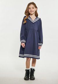 Navyblaues Kleid mit großem weiß gestreiften Matrosenkragen, langen Ärmeln und gerafft Taillierung. Kombiniert mit schwarzen Stiefeletten und weißen Socken.