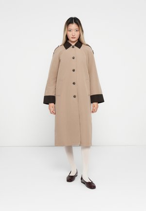 Woman standing, wearing a beige long coat with dark brown collar and cuffs, white tights, and dark red ballet flats against a plain background.