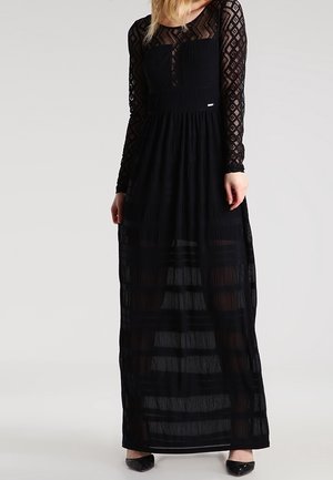 Femme portant une longue robe noire avec des manches longues en dentelle transparente et une jupe rayée transparente, se tenant devant un fond uni.