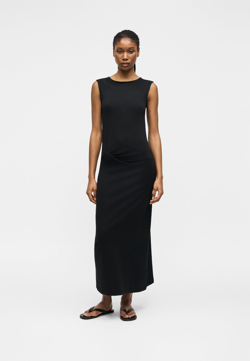 Γυναίκα όρθια, φοράει μια μαύρη μάξι αμάνικη φόρεμα και μαύρα σαγιονάρες σε απλό ανοιχτό γκρι φόντο.