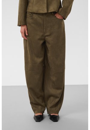 Personne portant un pantalon olive ample et une veste assortie, une main dans la poche et des chaussures noires pointues sur un sol gris.