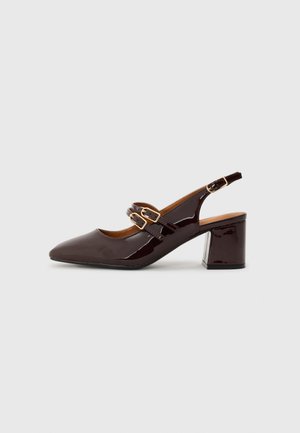 Temno rjave lakirane slingback čevlje z dvema zlatima zaponkama in blok peto na svetli podlagi.