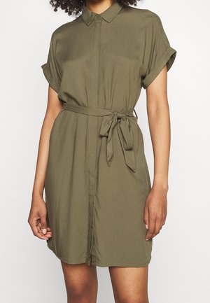 Kvinde iført en olivengrøn kortærmet skjortekjole med krave og et bundet bælte i taljen, stående foran en ensfarvet hvid baggrund.