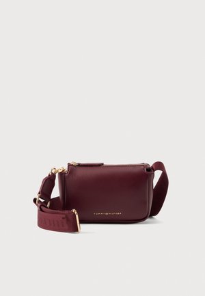 Sac bandoulière Tommy Hilfiger bordeaux, doté d’une large bandoulière réglable, de détails métalliques dorés et d’une fermeture zippée, sur fond clair.