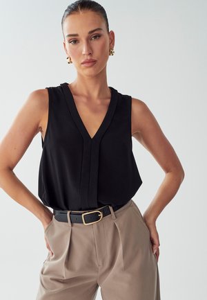 Top sans manches noir avec un décolleté en V profond, design drapé et détails plissés, associé à un pantalon beige taille haute et une ceinture noire.