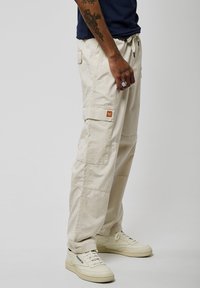 Pantalons cargo beiges avec plusieurs poches et une taille élastique, fabriqués à partir d'un tissu léger. Associés à des baskets blanches.