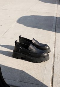 Ζευγάρι μαύρα χοντρά loafers με παχιά σόλα με πέλμα τοποθετημένα σε ανοιχτό γκρι πεζοδρόμιο με σκιές κοντά.