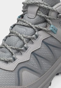 Κοντινό πλάνο γκρι αθλητικού παπουτσιού με ύφασμα από δίχτυ, κορδόνια με σχέδιο, συνθετικά επικαλύμματα και ανοιχτό μπλε διακριτικό κοντά στις τρύπες κορδονιών.