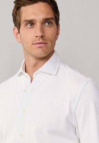 Młody mężczyzna z jasnobrązowymi włosami i zielonymi oczami, ubrany w białą koszulę zapinaną na guziki, patrzy lekko w lewo na jednolitym tle.