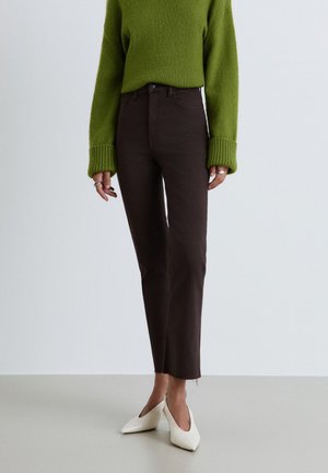 Persona che indossa un maglione verde, jeans marrone scuro a vita alta e gamba dritta, e tacchi bianchi a punta, in piedi su un pavimento grigio.