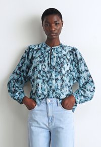 Blå och turkosmönstrad blus med puffärmar, plisserade detaljer och en nyckelhålskrage, tillsammans med ljus denimjeans.