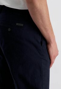Navy Baumwollmischung-Hosen mit einer Seitentasche, Gesäßtasche mit einem schwarzen Logo-Tag und einer glatten Textur. Sichtbares Knopfdetail auf der Rückseite.