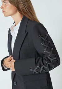 Črn blazer z natančno zasnovo, z okrasnim vezanjem in luknjami na rokavih. Narejen iz strukturirane tkanine, vključuje en žep na prsih.