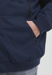Tamsiai mėlyna striukė su užtrauktuku, elastingais rankogaliais ir šonine kišene. Audinys yra lygus ir turi švelnų blizgesį.