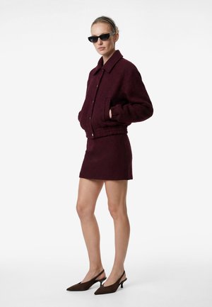 Femme portant une veste et une jupe bordeaux foncé, des lunettes de soleil noires forme œil de chat, un serre-tête noir et des talons slingback marron, debout les mains dans les poches.