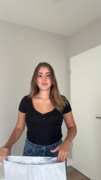 Top negru cu mânecă scurtă, cu decolteu rotund și detalii cu nasturi. Modelul ține un articol de îmbrăcăminte gri, pliat. Fundal deschis cu o ușă vizibilă.