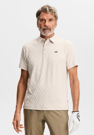 Mann mittleren Alters mit grauem Haar und Bart, der ein strukturiertes, cremeweißes Poloshirt, beige Hosen und einen weißen Golfhandschuh an der rechten Hand trägt.