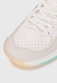 Hvit lær- og semsket skinnsneaker med perforerte detaljer, flate lisser og en gjennomsiktig gummisåle med en turkis aksent.