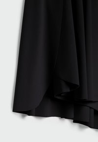 Tissu noir avec un ourlet superposé et asymétrique suspendu contre un fond blanc, montrant la texture et les détails de couture.