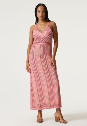 Femme aux cheveux tressés, portant une robe maxi sans manches à rayures roses et des sandales compensées ouvertes couleur beige, debout sur un fond uni.