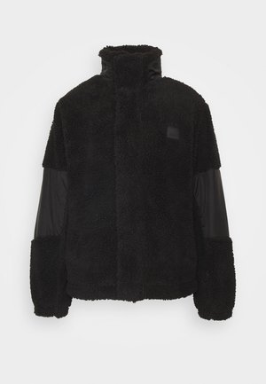 Giacca nera con corpo in fleece soffice e testurizzato, maniche in nylon liscio, collo alto e toppe con logo sul petto.