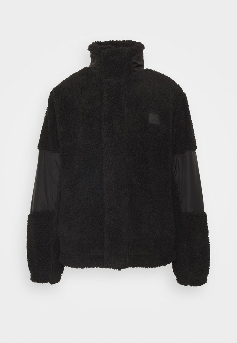 Sort jakke med en tekstureret, fluffy fleece-krop, glatte nylonærmer, høj krave og et logo-patch på brystet.