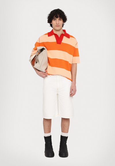 Gestreifter, orange und pfirsichfarbener Strickpolo mit rotem Kragen, kombiniert mit weißen Denim-Shorts und schwarzen Stiefeletten. Das Model hält eine beigefarbene Tasche.