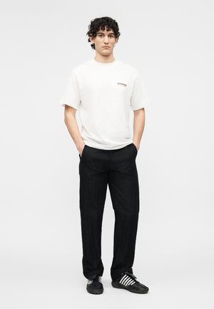 Ung mann med krøllete hår iført en hvit Barrow T-skjorte, svarte bukser og svarte Adidas-sneakers med hvite striper, hendene i lommer.