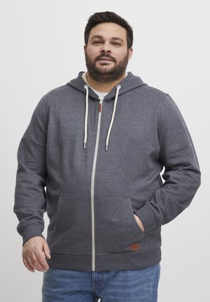 Siva zip-up dukserica izrađena od mekog materijala, sa prednjim zatvaračem, velikim džepovima, kapuljačom sa vezicom i kožnim detaljem na rubu.