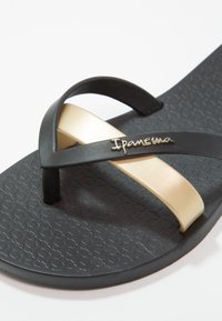 Tongs noires avec une lanière en accent doré, dotées d'une semelle texturée et d'un logo de marque "Ipanema" sur la lanière. Design plat.