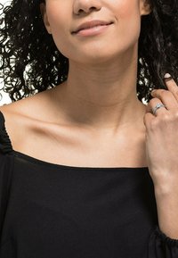 Donna con capelli ricci che indossa un top nero a spalle scoperte, mentre tiene delicatamente un dito con un anello d'argento con una pietra trasparente.
