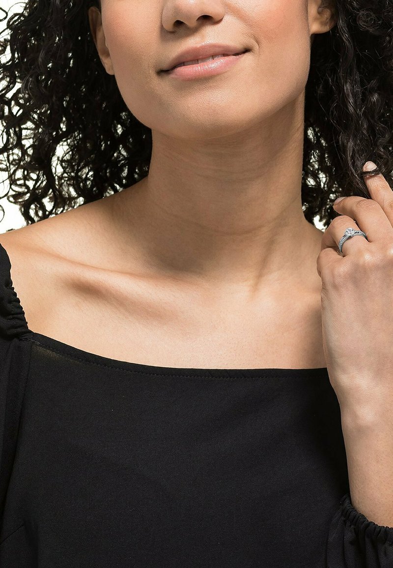 Kvinde med krøllet hår iført en sort off-shoulder top, der forsigtigt holder en finger med en sølvring prydet med en klar sten.