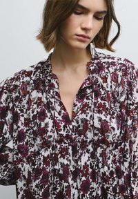 Blusa con motivo floreale bianca con accenti rosso scuro e viola. Presenta scollo a v, dettagli plissettati e tessuto texturizzato.