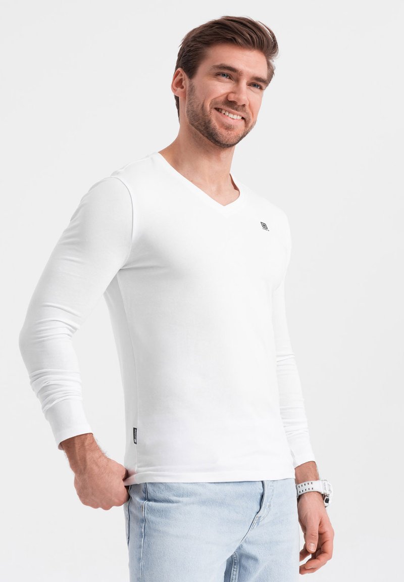 T-shirt bianco a maniche lunghe con scollo a V, in morbido cotone, design minimalista, presenta un piccolo logo sul petto, abbinato a jeans azzurri chiari.