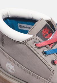 Stivale in pelle grigia con un colletto imbottito bianco, lacci rossi e blu, occhielli esagonali e un logo ad albero. Fodera interna a motivo blu e bianco.