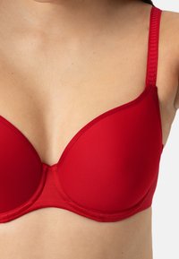 Punottu punainen rintaliivi, jossa on sileä kangas, säädettävät olkaimet ja kaareva muotoilu, korostaen saumattomasti muotoiltua siluettia ilman koristeluja.