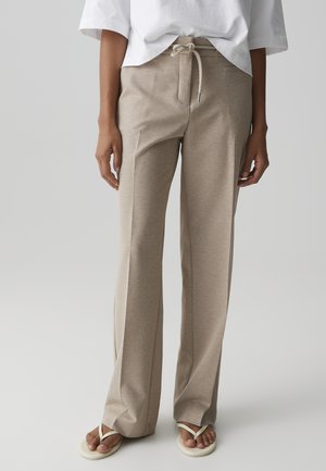 Persona che indossa pantaloni beige a gamba larga con vita con coulisse, top bianco a maniche corte e sandali infradito bianchi, in piedi contro uno sfondo neutro.