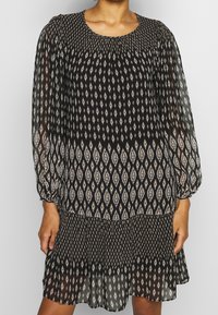 Svart långärmad klänning i lätt, genomskinligt tyg med ett diamantmönster i beige och vitt, med samlad halsringning och lager-på-lager nederdel.