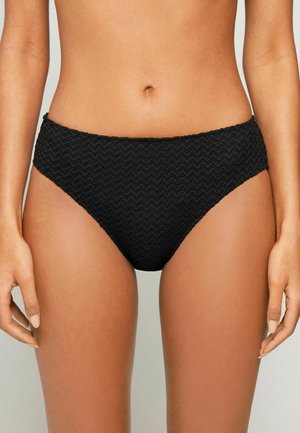 Frau trägt schwarze, texturierte High-Cut-Bikinihosen, zeigt Taille und obere Oberschenkel vor neutralem Hintergrund.