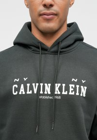 Felpa grigia scura realizzata in tessuto morbido, dotata di tasca a canguro, cappuccio con cordino e logo bianco "CALVIN KLEIN" con data di fondazione.