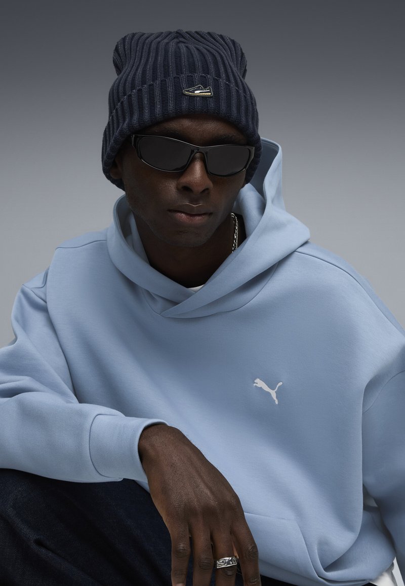 Sudadera azul claro con un logo blanco de Puma, combinada con un gorro de canalé azul marino y gafas de sol negras. Jeans de mezclilla oscura y un anillo plateado visible.