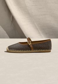 Καφέ επίπεδη εσπαντρίγια με μοτίβο μονόγραμμα, καφέ λουρί με χρυσή πόρπη και πλεκτή σόλα από γιούτα πάνω σε φωτεινή επιφάνεια και φόντο από σκιασμένο ύφασμα.