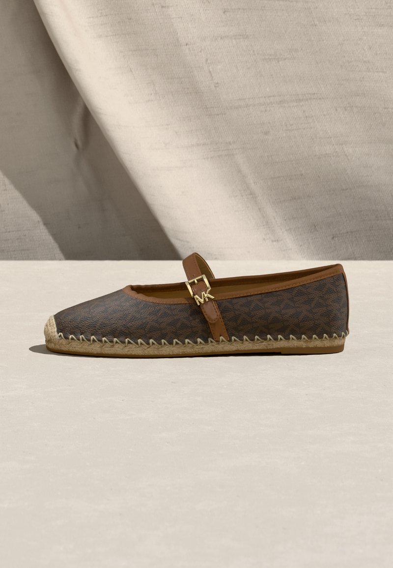 Καφέ επίπεδη εσπαντρίγια με μοτίβο μονόγραμμα, καφέ λουρί με χρυσή πόρπη και πλεκτή σόλα από γιούτα πάνω σε φωτεινή επιφάνεια και φόντο από σκιασμένο ύφασμα.