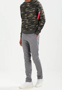 Felpa camouflage con orlo e polsini a coste; pantaloni grigi; sneaker bianche; tasca con zip e etichetta rossa sulla manica sinistra.