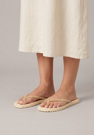 Lichtbeige geweven teenslippers gedragen op blote voeten met een crèmekleurige midi-rok zichtbaar boven de enkels op een neutrale achtergrond.