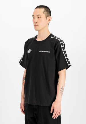 Mann mit kurzen Haaren steht und trägt ein schwarzes Alpha Industries T-Shirt mit weißem Logo und gemusterten Streifen auf den Schultern vor einem schlichten weißen Hintergrund.