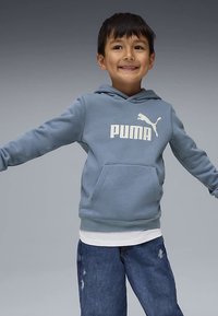 Ragazzo giovane e sorridente con capelli scuri, che indossa un felpa blu Puma, una maglietta bianca sotto e jeans blu, con le braccia aperte su uno sfondo grigio.