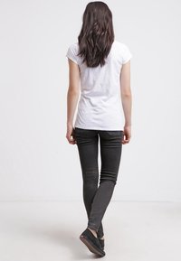 Weißes, eng anliegendes T-Shirt, schwarze Skinny-Jeans mit subtilen Details und dunkle Slipper. Modell, das sich abwendet, mit langen, sichtbaren Haaren.
