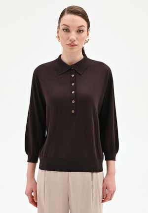 Donna che indossa un maglione polo marrone scuro a maniche lunghe con bottoni e pantaloni beige a gamba larga, in piedi su uno sfondo bianco.