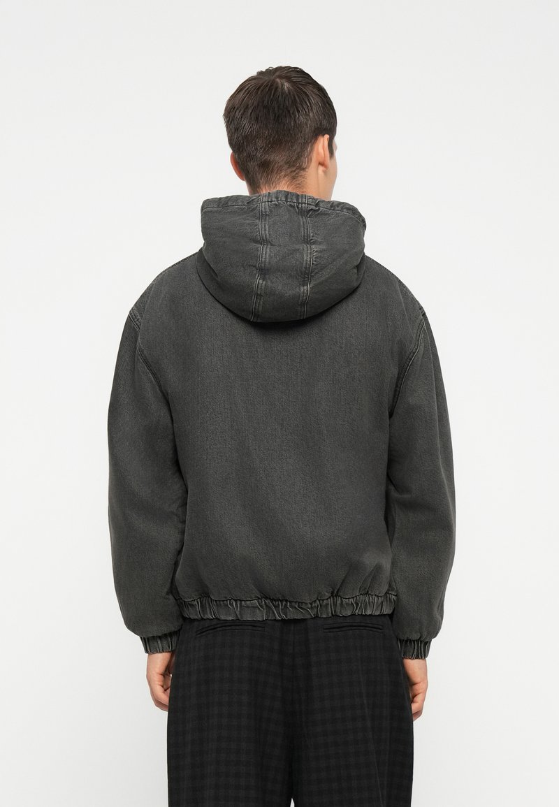 Chaqueta con capucha de color gris oscuro, con puños y cintura elásticos, fabricada en tela de mezclilla. Presenta un corte relajado y detalles de costura visibles.